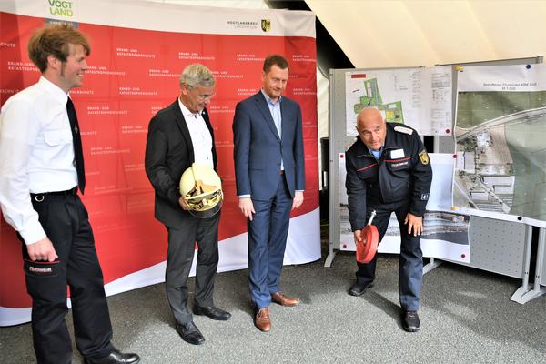 Der Vorsitzende des Kreisfeuerwehrverbandes Vogtland e.V., Andreas Schubert zeigt vor Ministerpr&auml;sident Michael Kretschmer, Landrat Rolf Keil und Kreisbrandmeister Gerd P&uuml;rzel, wie man einen Feuerwehrschlauch ausrollt. Landrat Rolf Keil h&auml;lt einen Feuerwehrhelm in der Hand.